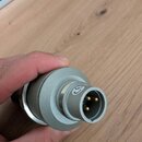 Detailní záběr na kovový konektor stříbrné barvy s několika zlatými piny uvnitř. Na povrchu je vyryto logo připomínající stylizované písmeno C. Povrc…