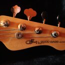Fotografie zachycuje hlavu basové kytary z přírodního dřeva se čtyřmi kovovými ladičkami. Na dřevě je černý nápis „G&L L-2000 BASS SERIES E“ a pod ní…