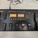 Černý kytarový procesor s nápisem BOSS GT-100 COSM AMP EFFECTS PROCESSOR. Zařízení má displej s oranžovým textem BOSS GT-100 Ver. 2, několik ovládací…