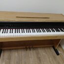 Digitální piano v dřevěné kostelní konstrukci světle hnědé barvy. Na černém ovládacím panelu je vpravo nápis "Roland". Nástroj má standardní klávesni…