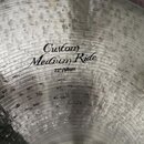 Detailní pohled na kovový činel s viditelným kovaným povrchem. Uprostřed je černý nápis „Custom Medium Ride“ a pod ním označení „22"/56cm“. Povrch vy…