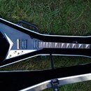 Černá elektrická kytara s tělem ve tvaru V v otevřeném černém kufru. Na hlavě hředu je bílé logo Jackson. Kytara má dva humbuckery, zlatý most a ladí…