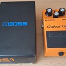 Kytarový efekt v oranžovém kovovém provedení s černým pedálem. Na zařízení jsou nápisy Distortion, DS-1 a logo BOSS. Vedle efektu je černá krabice s…
