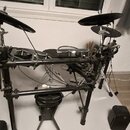 Elektronická bicí souprava s černým kovovým rámem a gumovými pady. Souprava obsahuje dva činely, snare drum, tom-tomy a basový pedál. Na komponentech…