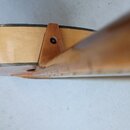 Detailní pohled na spoj hnedebního nástroje, pravděpodobně kytary, kde je krk spojen s korpusem. Nástroj je vyroben ze světlého dřeva s tmavým lemová…