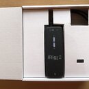 Černý obdélníkový modul iRig PRE 2 umístěný v bílém kartonovém obalu. Na přední straně jsou nápisy 48V, ON a logo iRig PRE 2. V horní části jsou ikon…