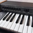 Detailní pohled na klávesový nástroj s černým povrchem a dřevěnými bočními listy. Na ovládacím panelu jsou nápisy PERCUSSION (s tlačítky DIRECT, SLOW…