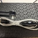 Stříbrný opletený kabel s černým plastovým převodníkem. Na převodníku jsou bílé nápisy MIDI, IN, OUT a USB spolu s logem připomínajícím stylizedovano…