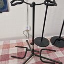 Černý kovový stojan na kytaru s gumovými nástavci v horní části a spodní oporou. Stojan stojí na červenobílém kostkovaném ubrusu. Na povrchu jsou vid…