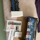 Na fotografii je modrý kytarový procesor s nápisem BEHRINGER V-AMP PRO, umístěný v polystyrenovém obalu. Vedle něj je otevřená kartonová krabice s pr…