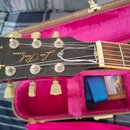 Detail hlavy černého elektrického kytary s nápisy Gibson, Les Paul a CLASSIC. Nástroj má stříbrné ladičky s krémovými knoflíky. Kytara leží v otevřen…