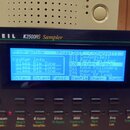 Černý sampler s nápisem KURZWEIL K2500RS Sampler. Zařízení má modrý podsvícený displej, na kterém je zobrazen text ProgramMode, XPose:368T a Channel:…