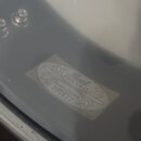 Detail bílé membrány bubnu s oválnou nálepkou. Na nálepce je nápis Pearl, PICCOLO a Snare Drum s číslem 200613. Povrch membrány vykazuje drobné nečis…