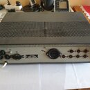 Šedý elektronický přístroj v kovovém šasi s perforovaným horním krytem. Na zadním panelu jsou umístěny konektory, přepínače a štítky s nápisy 16A/220…