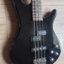 Černá elektrická basová kytara s tmavým hmatníkem. Nástroj má dva snímače typu humbucker a tři ovládací knoflíky. Povrch těla vykazuje drobné škrában…