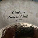 Detailní záběr na povrch činchy z kovu. Na povrchu je černý nápis „Custom Hybrid Crash“ a pod ním označení „19"/48cm“. Levá část fotky ukazuje kovaný…