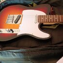 Elektrická kytara s tělem v červeno-žlutém sunburst provedení a bílým pickguardem. Nástroj má hnědý hриф s dřevěnou strukturou a kovový most s integr…