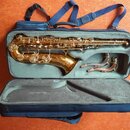 Zlatý saxofon v modrém měkkém pouzdře s šedým vnitřkem. Vedle nástroje je v pouzdře umístěn zahnutý hnítek. Na instrumentu nejsou viditelné žádné zna…