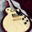 Krémová elektrická kytara s tvarem single-cut v otevřeném kufru s červeným polstrováním. Nástroj má dva humbuckery se zlatými kryty, zlatý kobylník a…