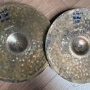 Dva kovyvové činely s kovaným povrchem. Na levém je modrý nápis „35 cm 14 Hi-Hat BOTTOM“ a bílý nápis „MADE IN TURKEY“, v centru je viditelné číslo „…