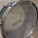 Fotografie znázorňuje snare drum s průhlednou membránou a kovovým obručem. Na membráně je vidět nápis „EVANS“ a logo značky „Remo“. Přes membránu je…
