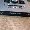 Šedé audio zařízení s ovládacími prvky na horní straně, včetně velkého otočného knoflíka a řady tlačítek. Na předním černém panelu je bílý nápis UNIV…