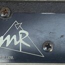 Detail zadní strany černého elektronického zařízení s napájecím konektorem a třemi MIDI porty. Na povrchu je bílé logo tvořené trojúhelníkem a styliz…