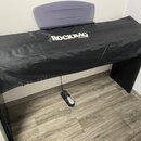 Černý krycí potah z syntetického materiálu zakrývá klavír nebo digitální piano. Na horní části potahu je bílý nápis ROCKBAG by Warwick. Pod nástrojem…
