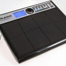 Černý elektronický bicí pad s osmi gumovými polštářky. V horní části je stříbrný panel s modrým displejem, ovládacím knoflíkem a řadou tlačítek. Na p…