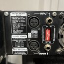 Detail zadní strany černého elektronického zařízení s dvěma vstupy. Jsou zde viditelné konektory XLR a jack, označené jako INPUT 1 a INPUT 2. Na přís…