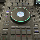 Černý DJ controller s nápisem DENON DJ. Zařízení obsahuje velký jogwheel s podsvícením, několik posuvníků, otočných knoflíků a modře podświetlených t…
