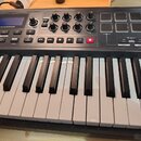 Černý MIDI kontrolér s klávesnicí. Na horním panelu je displej s nápisem novation, osm otočných potenciometrů a osm čtvercových padů. Jsou zde vidite…