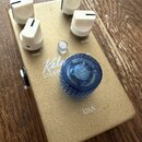 Zlatý kytarový efekt s texturou metalického povrchu. Na horní straně jsou čtyři bílé plastové otočné knoflíky s nápisy TONE a GLASS, jeden malý stříb…