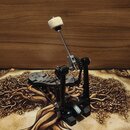 Fotografie znázorňuje černý basový pedál s kovovými komponenty a řetězovým pohonem. Beater je zakončen bílým plstěným hrotem. Na šlapadle jsou vidět…