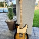 Elektrická kytara s tělem v provedení sunburst a bílým pickguardem. Na hlavě hředu je viditelné logo Fender. Kytara má světle dřevěný hmatník a zlaté…