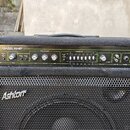 Černý basový kombo zesilovač s kovovou mřížkou reproduktoru. Na horním ovládacím panelu je nápis BA150 BASS AMP a logo Ashton. Na mřížce je bílý nápi…