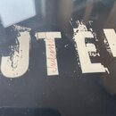 Detailní záběr na černý povrch s bílým nápisem. Jsou vidět části písmen „JTE“ a v mezích je svisle napsáno slovo „Julion“. Nátěr nápisu vykazuje výra…