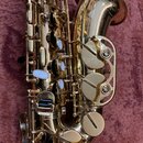 Zlatý saxofon v otevřeném kufru s fialovým vnitřkem. Na nástroji jsou vidět bílé podložky klavíů a kovové mechanismy. Povrch je lesklý, s viditelnými…