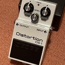 Bílý efekt pedál s nápisy Distortion a DS-1. Na horní části jsou tři ovládací knoflíky s popisky LEVEL, TONE a DIST a červená kontrolka s nápisem CHE…