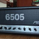 Kytarový zesilovač s černým povrchem a kovovou mřížkou. Na mřížce je bílý nápis 6505 a logo PEAVEY. Spodní ovládací panel obsahuje různé potenciometr…