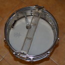 Fotografie zachicuje pohled shora na snare drum s bílým blátcem. Na blátku je vidět logo EVANS a nečitelné kulaté logo s nápisem. Přes buben je polož…
