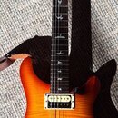 Elektrická kytara s žlutě-oranžovým přechodem (sunburst) a viditnou strukturou dřeva. Na hlavě je červený lak a bílý nápis PRS. Hmatník má vložky ve…