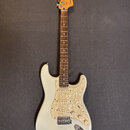 Elektrická kytara s bílým trupem a hmatníkem z tmavého dřeva. Na hlavě je nápis Fender STRATOCASTER. Kytara má tři snímače a plech s perleťovým vzore…