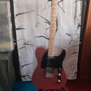 Elektrická kytara s růžovým tělem a černým pickguardem umístěná na stojanu. Na hlavě hředu je vidět nápis Fender TELECASTER. Hmatovnice a krk jsou vy…