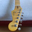 Fotografie zachicuje hlavu elektrické kytary v přirozené dřevěné barvě. Na hlavě je vidět logo Fender® a nápis STRATOCASTER®. V horní části loga je u…