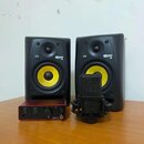 Dva černé studiové monitory s žlutými membránami. Na obou jsou viditelná loga KRK a nápisy ROKIT 5. Před nimi stojí červený zvukový interface s nápis…