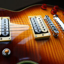Detail elektrické kytary s tělem v žlutém flame-maple provedení a červeným okrajem. Nástroj má dva humbuckery s krémovými rámečky, chromovaný kobylní…