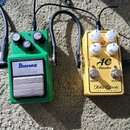 Dvě kytarové efektní pedály položené na koberci. Vlevo je zelený pedal s nápisem Ibanez TS9 TubeScreamer a vpravo žlutý pedal s nápisy AC-booster, Xo…