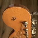 Záběr na zadní stranu hlavy kytary z přírodního dřeva s kovovými ladičkami. Na dřevě jsou čitelné nápisy „ORIGINAL Contour Body“ a „STRATOCASTER“, dá…