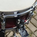 Detailní pohled na červený buben s chromovaným obručem a bílým koženem. Na korpusu je připevněn chromovaný držák s černým plastovým knoflíkem. Na spo…